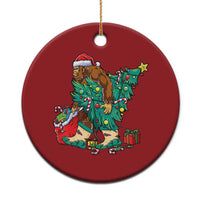 Red round holiday ornament with a cartoon Bigfoot Santa decorating a Christmas tree.