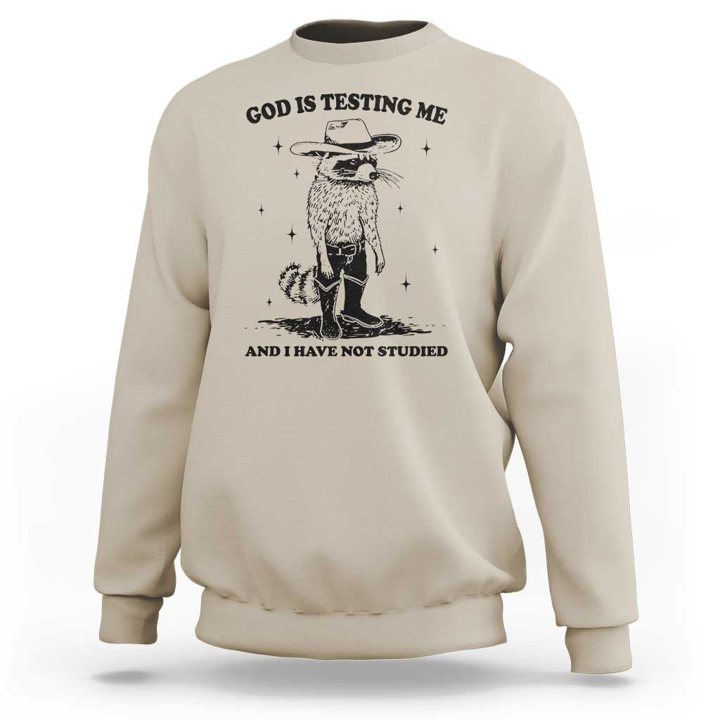 Beige sweatshirt with cowboy raccoon graphic and "God is testing me" text