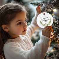 Personalized engaged circle Christmas ornament held near a tree, with “Engaged,” James & Emily, 2024.