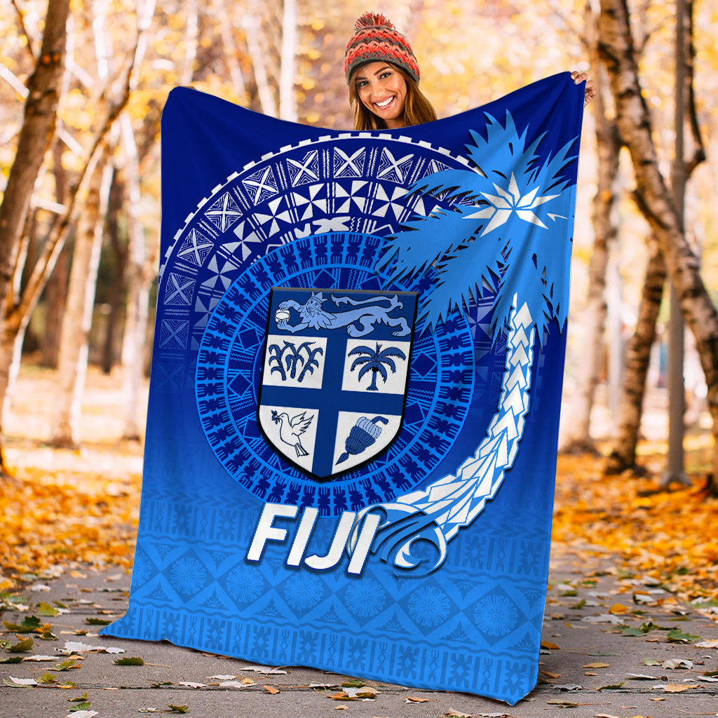 Blue Fiji tapa blanket held outdoors, featuring shield crest and “FIJI” lettering