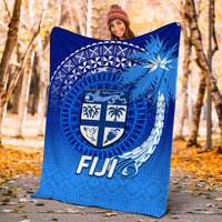 Blue Fiji tapa blanket held outdoors, featuring shield crest and “FIJI” lettering