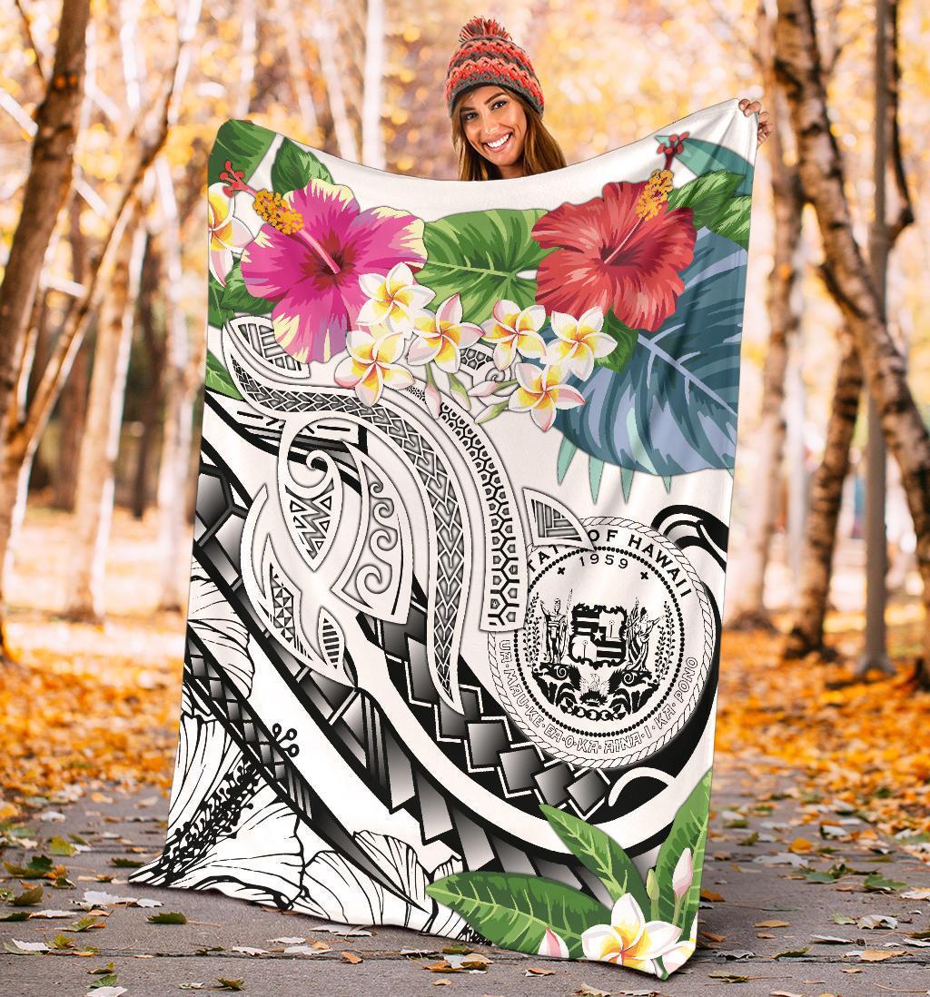 White Polynesian blanket held outdoors with plumeria blossoms, tropical leaves, and tribal emblem