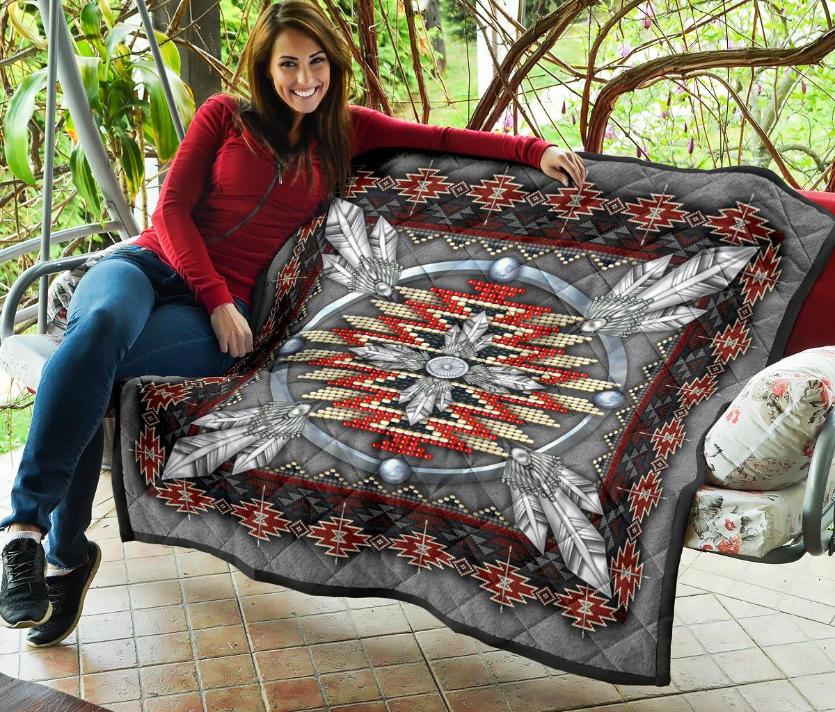 Grey quilt draped over chair, showing feather medallion with red/white beadwork and zigzag trim.