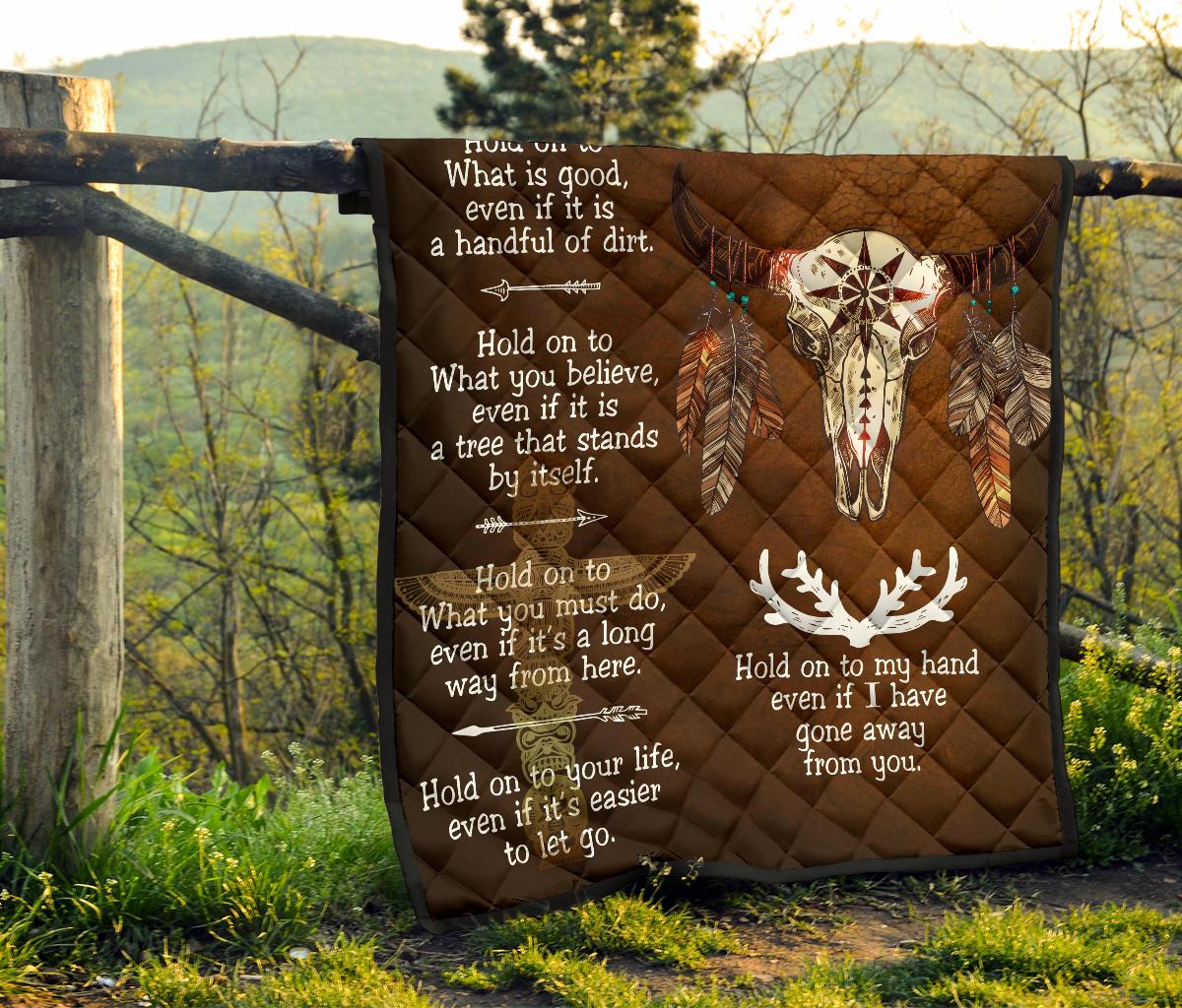 Brown quilt hanging on fence post with bison head, feather details, and white quotes